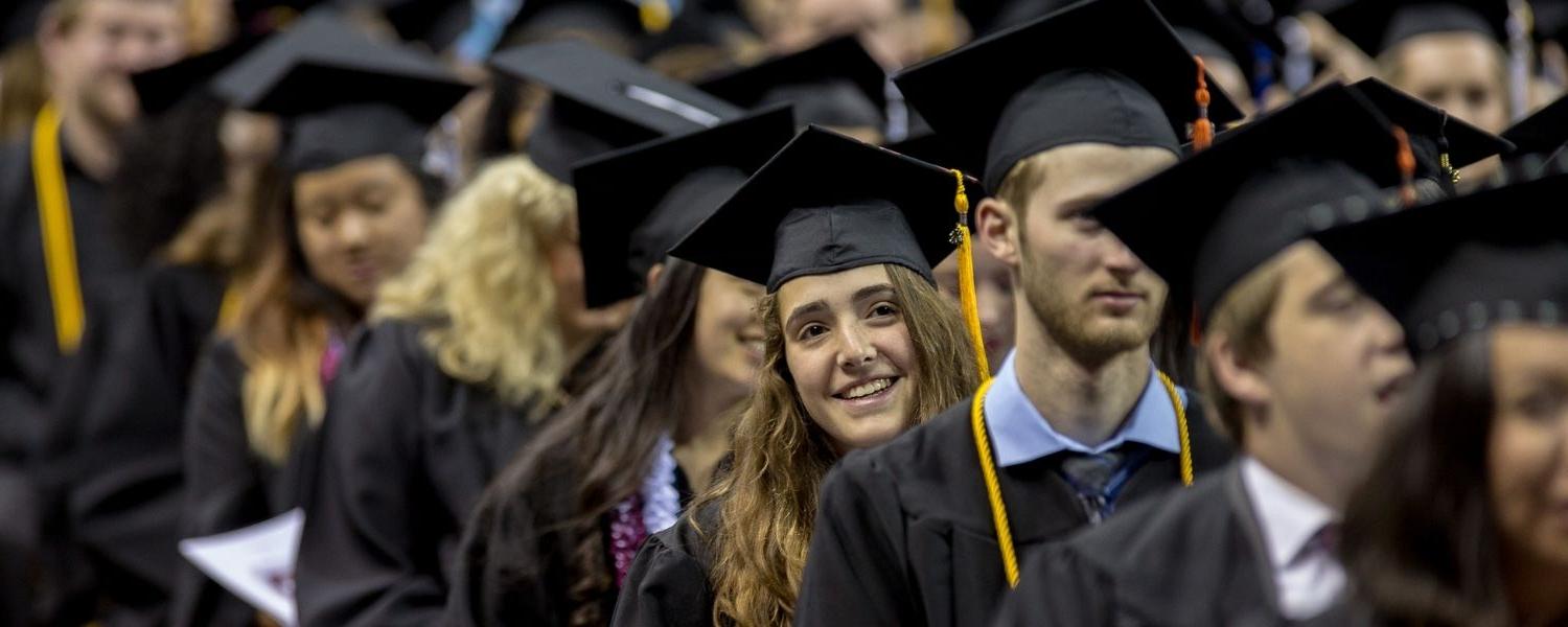 SPU graduates line up to begin the Commencement ceremony; one young graduate leans to her left, 微笑, 朝舞台前面看.